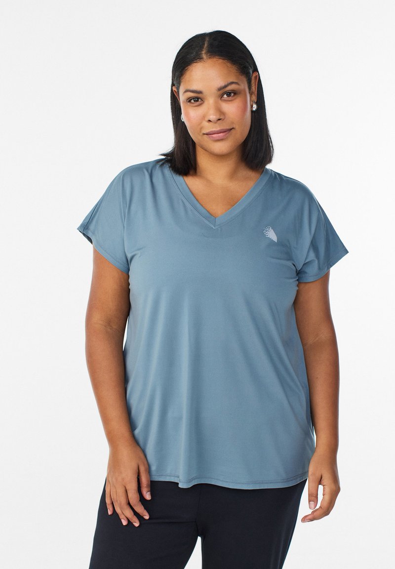 Femme aux cheveux noirs lisses portant un T-shirt bleu à col en V et un pantalon noir, debout devant un fond blanc uni.