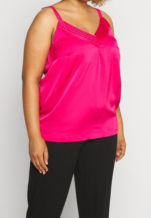 Person iført en lys pink ærmeløs satinbluse med plisseret V-hals og sorte bukser, der står foran en ensfarvet baggrund.