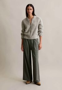 Egy textúrált, szürke cipzáras pulóvert viselő nő áll egy sima háttér előtt, széles lábú kockás nadrágban és világosbarna hegyes orrú cipőkben.