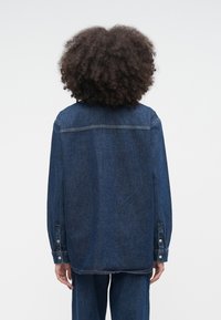 Osoba o kręconych włosach, ubrana w luźną, ciemnoniebieską kurtkę dżinsową i dopasowane spodnie, stoi tyłem na jednolitym jasnym tle.