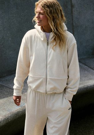 Vrouw met lang krullend haar die een witte hoodie met rits en bijpassende joggingbroek draagt, staand buiten bij een stenen bank.