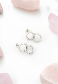 Silverfärgade hoopsörhängen med sammanflätade cirkelformer. Slät, polerad metall med en glänsande yta, mot en ljus bakgrund.