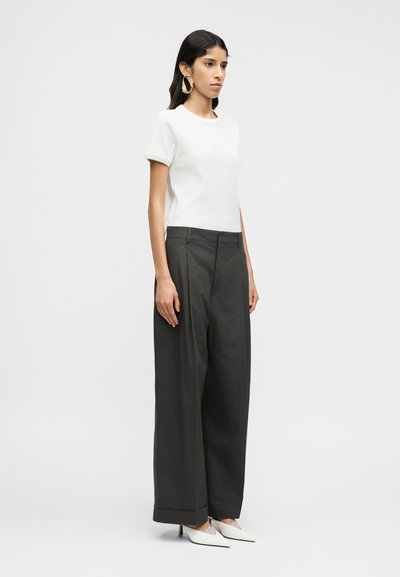 Λευκό τοπ με κοντά μανίκια, γκρι παντελόνι ψηλόμεσο με φαρδιά μπατζάκια και λευκά ψηλοτάκουνα παπούτσια με μυτερή μύτη και δαντελένιες λεπτομέρειες, φορεμένα από μοντέλο που στέκεται.