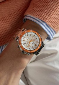 Sviandestål klocka i silverfärg med en orange bezel, vit urtavla och orange detaljer. Innehåller kronograf sub-dialer och en metallarmband.