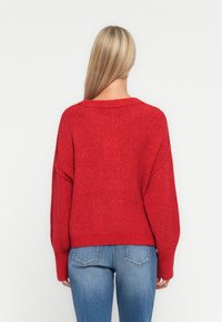 Un pull rouge avec une coupe ample et une texture côtelée, doté d'un large décolleté et d'épaules tombantes, associé à un jean bleu.