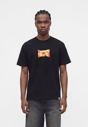 Jonge man met gevlochten haar die een zwart T-shirt draagt met een kleurrijke snackverpakkingsafbeelding in het midden, gecombineerd met donkergrijze spijkerbroek.