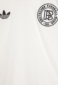 Witte rib jersey met het Adidas-logo en een cirkelvormig zwart embleem met "Deutscher Fußball-Bund" en "125." Subtiele textuurdetail.