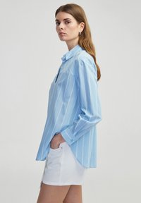 Camisa de manga larga azul claro con rayas blancas verticales, cuello abotonado y aberturas laterales. Combinada con pantalones cortos de mezclilla blancos.