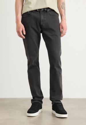 Person wearing black slim-fit jeans, black sneakers with white soles, and a light beige shirt, standing on a light floor against a plain wall.