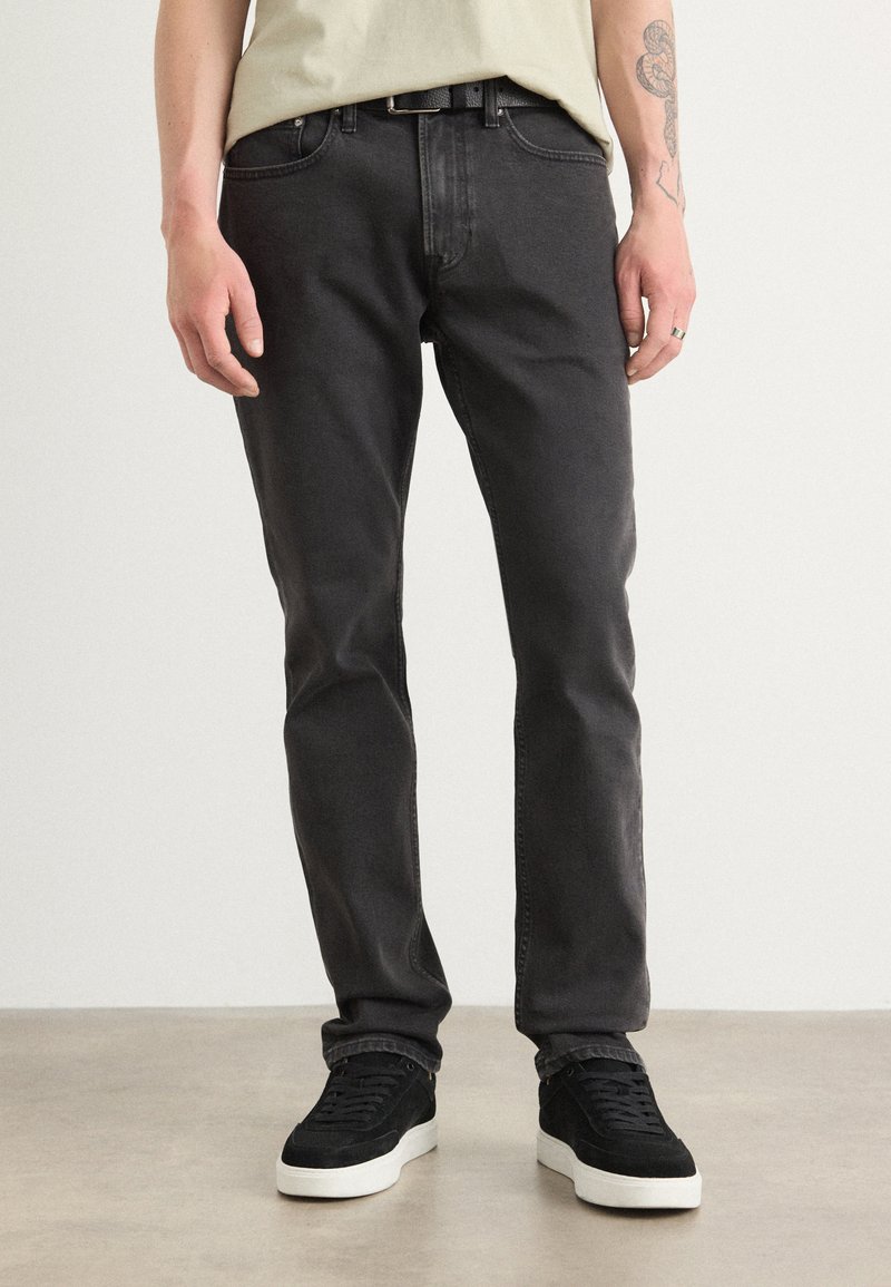 Persona che indossa jeans neri slim fit, sneakers nere con suola bianca e una camicia beige chiaro, in piedi su un pavimento chiaro contro un muro semplice.