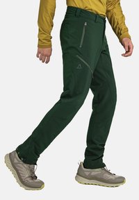 Grüne Outdoor-Hosen mit figurbetontem Design, ausgestattet mit Reißverschlusstaschen und einem strukturierten Stoff. Kombiniert mit hellgrauen Schuhen.