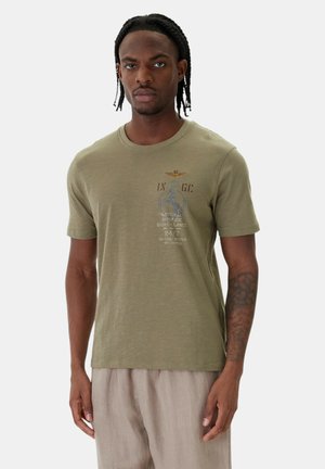Giovane uomo con capelli intrecciati che indossa una t-shirt grafica verde oliva e pantaloni beige, in piedi davanti a uno sfondo semplice.