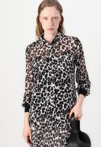 Leopardenmuster durchsichtige Button-Up-Bluse mit schwarzen Akzentmanschetten, zwei Fronttaschen und einer leichten, fließenden Textur.