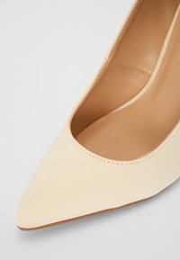 Scarpa con tacco alta beige, con punta affusolata, tomaia in sintetico liscia e design minimalista. L'interno presenta una fodera di un leggero color tan.
