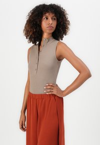 Tan ribbed sleeveless bodysuit with a high collar and visible button closure, paired with loose orange pants. Soft texture and relaxed fit.