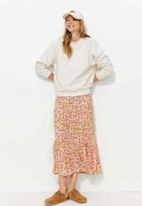 Sweatshirt beige clair, jupe à volants florale avec des motifs roses et oranges, chaussures slip-on marron, et casquette blanche.