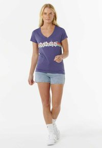 Paars v-hals t-shirt met bloemenpatroon en horizontale strepen over de borst, gecombineerd met lichte denim shorts en witte sneakers.