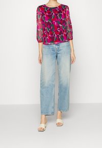 Haut transparent violet à motif floral, manches trois-quarts, associé à un jean large bleu clair et des sandales crème à enfiler.