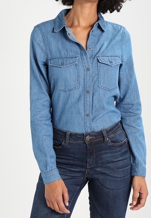 Person iført en langærmet denim skjorte med klaplommer, stoppet ned i mørkeblå denimjeans.