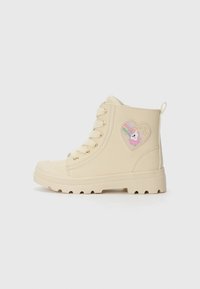 Botas de tobillo beige con ojales dorados, suela de goma texturizada y un diseño de unicornio en forma de corazón en el lado exterior.