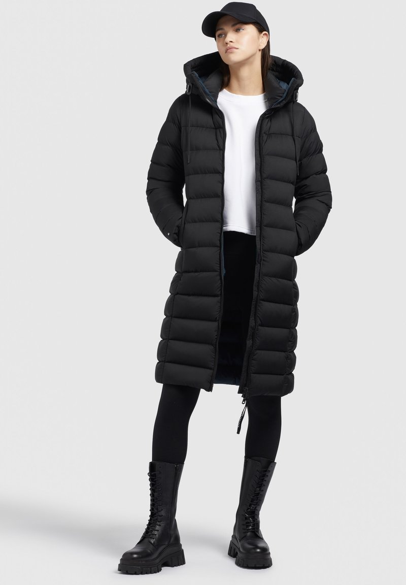 Schwarzer gepolsterter langer Mantel mit Reißverschluss vorne und Kapuze, ausgestattet mit einem gesteppten Design. Getragen über einem weißen Hemd und kombiniert mit schwarzen Leggings und Stiefeln.