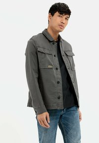 Grå denimjakke med knaplukning, to frontlommer og krave, stylet over en sort skjorte, parret med blå jeans.