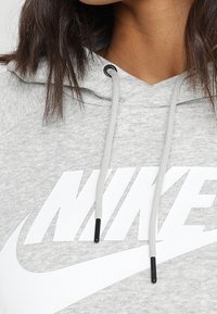 Šedá mikina s kapucí s velkým bílým logem Nike, kapucí se stahovacími šňůrkami s černými konci, měkkou texturou a volným střihem.