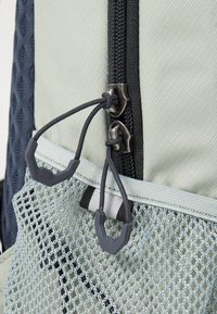 Texturiertes hellgrünes Rucksackmaterial mit dunkelblauem Netz, ausgestattet mit doppelten Reißverschlüssen, verstellbaren Schnüren und einem grauen Akzenttab.