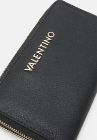 Svart teksturert lommebok med gull "VALENTINO"-logo, glidelås og sydd kanter. Har en glatt overflate og kompakt design.