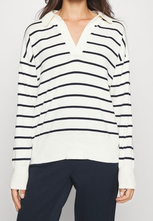 Vrouw draagt een wit shirt met lange mouwen en zwarte horizontale strepen, met een V-hals en een donkere broek.