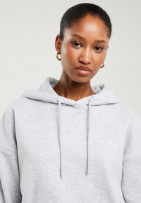 Młoda kobieta z krótkimi włosami, nosząca złote kolczyki-koła i jasnoszarą bluzę z kapturem ze ściągaczami na neutralnym tle.