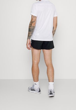 Mann mit Tätowierungen, der ein weißes Kurzarmhemd, schwarze Shorts, weiße Socken und graue Adidas-Sneaker trägt, steht vor einem einfarbigen Hintergrund.