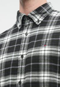Camisa de quadrados preto e branco com colarinho abotoado, apresentando botões pretos e um pequeno logótipo bordado em vermelho, branco e azul.