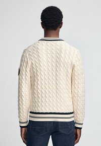 Kremowy sweter w warkoczowy splot z granatowymi prążkowanymi ściągaczami przy mankietach i u dołu. Posiada zaokrąglony kołnierz oraz naszywkę na lewym ramieniu.