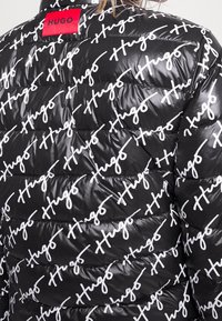 Sort pufferjakke med hvid skrifttype-logo printet over hele jakken. Rødt mærke med "HUGO" ved kraven. Tekstureret, skinnende stof og quiltet design.