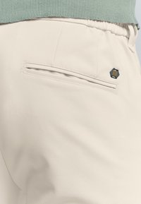 Beige broek van gestructureerde stof, met een achterzak en een zwart hexagonaal logo accent. Elastische tailleband zichtbaar aan de bovenkant.