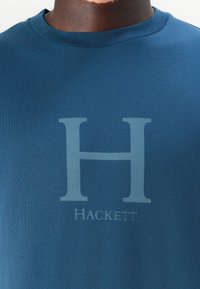 Tumši zila kokvilnas T-kreklā ar apaļu kaklu, uz kura ir liels gaiši pelēks "H" un zem tā izsmalcinātā dizainā ir vārds "HACKETT".