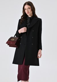 Cappotto in lana nera con design doppiopetto, grandi bottoni e rever a punta, abbinato a una gonna midi bordeaux e una borsa strutturata.