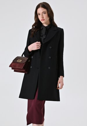 Cappotto in lana nera con design doppiopetto, grandi bottoni e rever a punta, abbinato a una gonna midi bordeaux e una borsa strutturata.