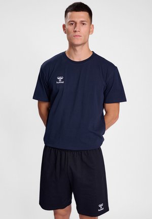 Mand står med hænderne bag ryggen iført navyblå Hummel sport t-shirt og shorts mod en ensfarvet lys baggrund.