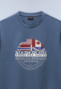T-shirt en coton bleu avec un graphisme rond comprenant les drapeaux des États-Unis, du Canada, de la Norvège et du Royaume-Uni, ainsi que le texte pour le Programme de Recherche Antarctique Napapijri.