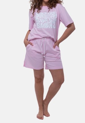 Vrouw draagt lichtroze shorts en bijpassend T-shirt met witte "SUNDAYS" tekst, staat blootsvoets met handen in de zakken.