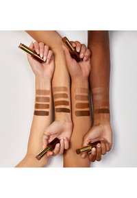 Vier armen met verschillende huidskleuren tonen make-up swatches in bruintinten. Elke arm houdt een bruine lipsticktube met een gouden accent vast.