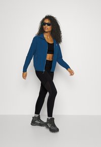 Blue zip-up jacket with long sleeves, black athletic top, black leggings with mesh detail, and black hiking shoes. Model stands on a light background.