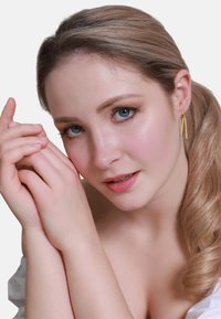 Nahaufnahme einer Person mit langen, hellbraunen Haaren, blauen Augen und goldenen abgehängten Ohrringen. Glatte Haut und sanftes, natürliches Makeup.
