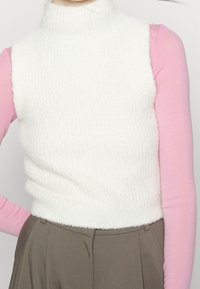 Top senza maniche white in tessuto a collo alto, sovrapposto a una camicia a maniche lunghe rosa, abbinato a pantaloni marroni a vita alta.