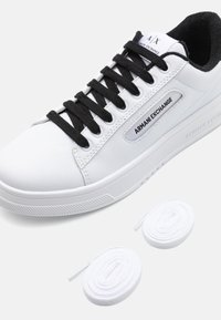 Bílé kožené tenisky s černými tkaničkami, s logem "Armani Exchange" na boku. Na zemi jsou dvě bílé tkaničky.