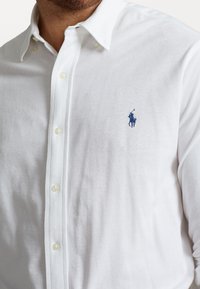 Man draagt een wit overhemd met knopen en een blauw geborduurd polospelerlogo op de linkerborst.