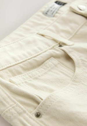 Beige Denim-Stoff mit strukturierter Oberfläche, ausgestattet mit einer Vordertasche, Knopfverschluss und kontrastierenden Nähten.