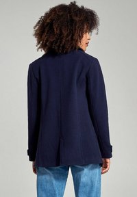 Cappotto blu navy in misto lana con una vestibilità su misura, spacco posteriore e polsini con bottoni, abbinato a denim azzurro chiaro. Texture morbida e design strutturato.
